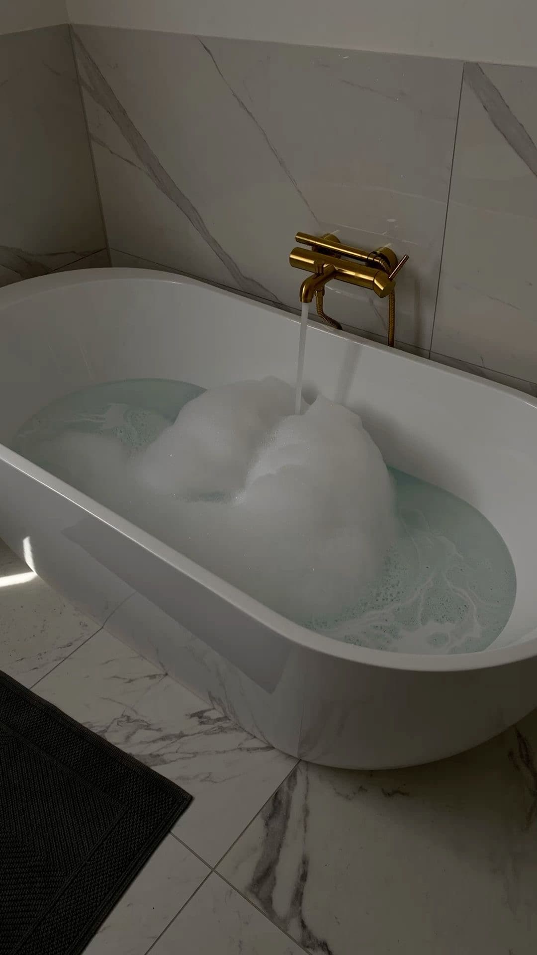 Freestanding bathtub with gold mixer tap and hand shower filling with water in a marble-tiled bathroom